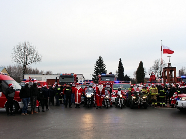 Na zdjęciu w tle samochody strażackie, z przodu strażacy w tradycyjnych strojach oraz mikołajowych czapkach. Pośrodku w towarzystwie motocyklistów stoi Św. Mikołaj.  Na wspólnym zdjęciu jest około 30 osób.