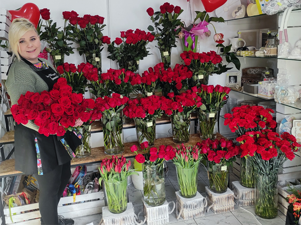 Na zdjęciu: Karolina Fyda - właścicielka Kwiaciarni Sasanka w Gorzycach.  Pani Karolina na rekach trzyma bukiet czerwonych róż, sfotografowana jest na tle kwiatów tj. czerwonych róż,  goździków i tulipanów, które ustawione są we flakonach na wystawie w kwiaciarni.