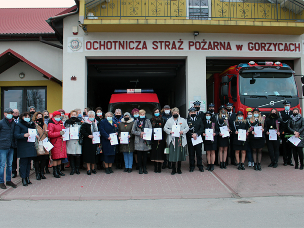 Na zdjęciu – przed budynkiem remizy OSP Gorzyce stoją druhowie strażacy, przedstawiciele samorządu oraz panie z KGW, stowarzyszeń i organizacji pozarządowych. Panie w rękach trzymają goździki i okolicznościowe adresy. 