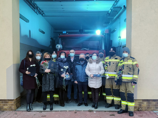 Na zdjęciu – przed budynkiem remizy OSP Trześń stoją druhowie strażacy, przedstawiciele samorządu oraz przedstawicielki  organizacji pozarządowych i stowarzyszeń. Panie w rękach trzymają goździki i okolicznościowe adresy. 