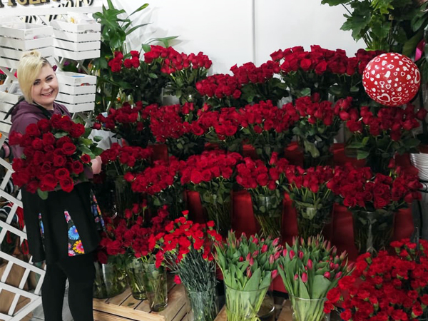 Wystawa w kwiaciarni, głównie z kwiatami w koloże czerwonym, przeważają róże. Oboj stoi kobieta, na ęku trzyma bukiet z czerwonych róż.